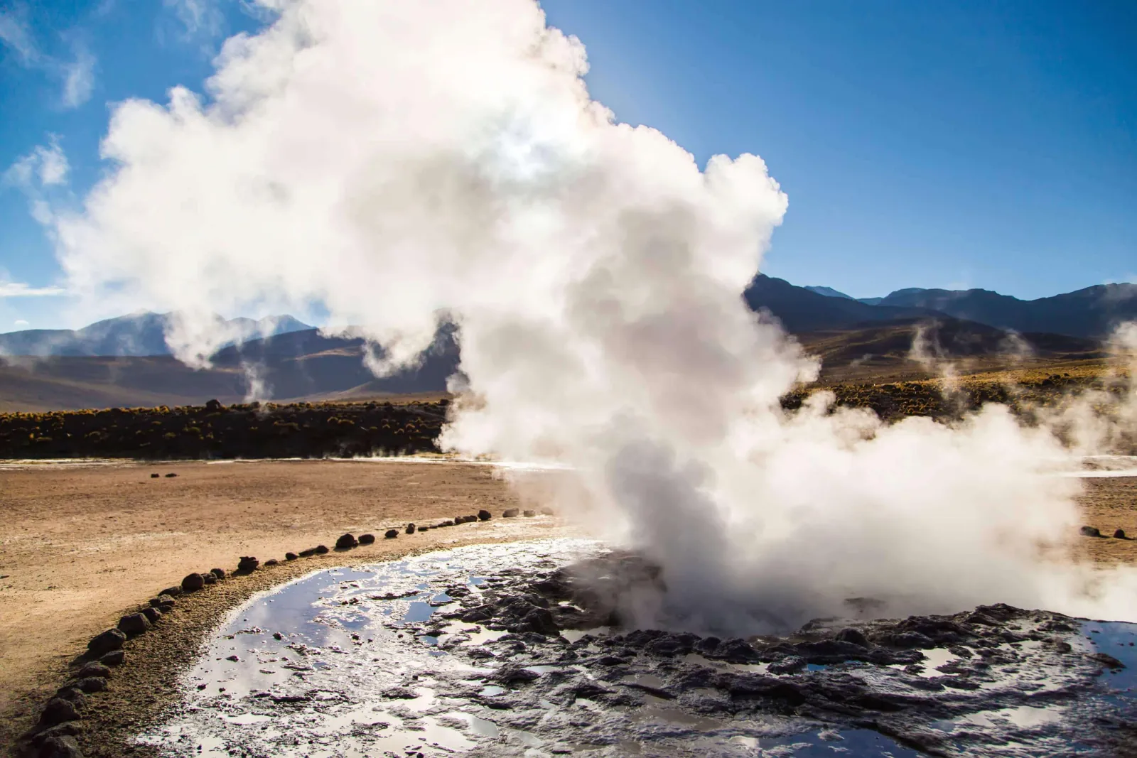 A tall column of steam rises from a geyser vent at El Tatio under a blue sky