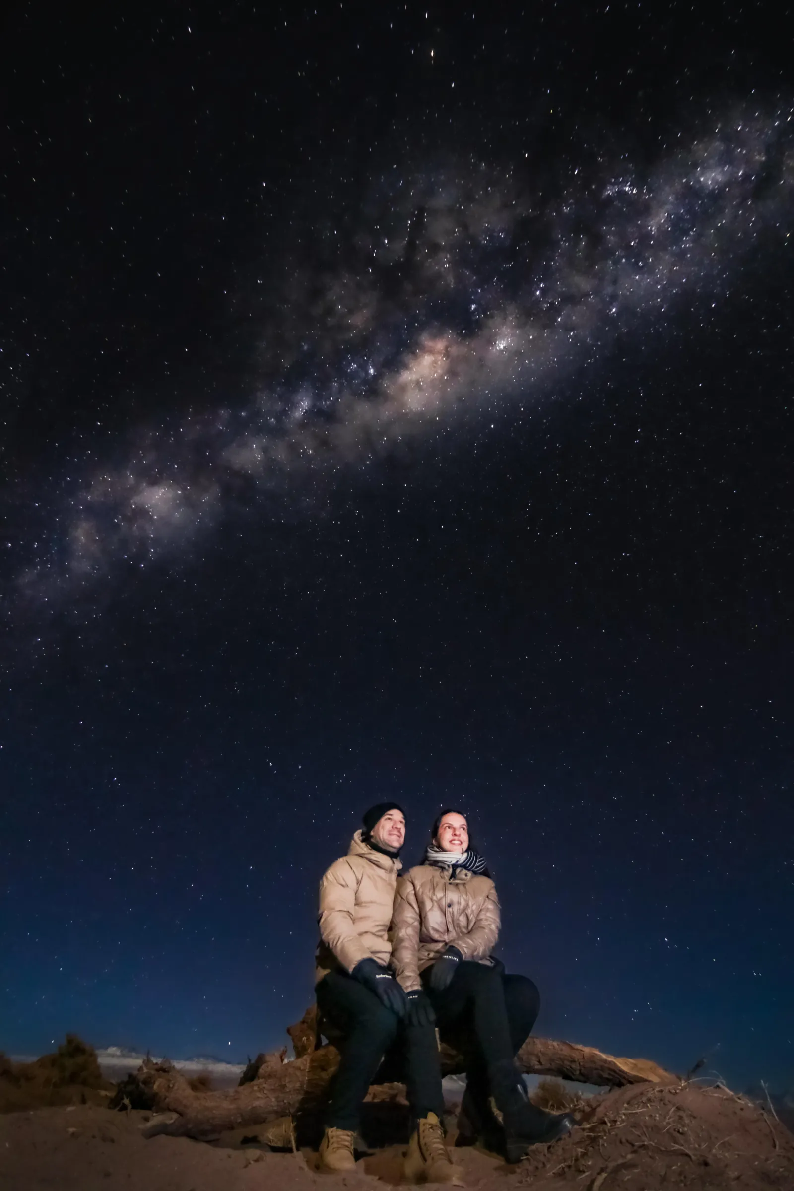 Two travellers in padded jackets sit side by side beneath the Milky Way