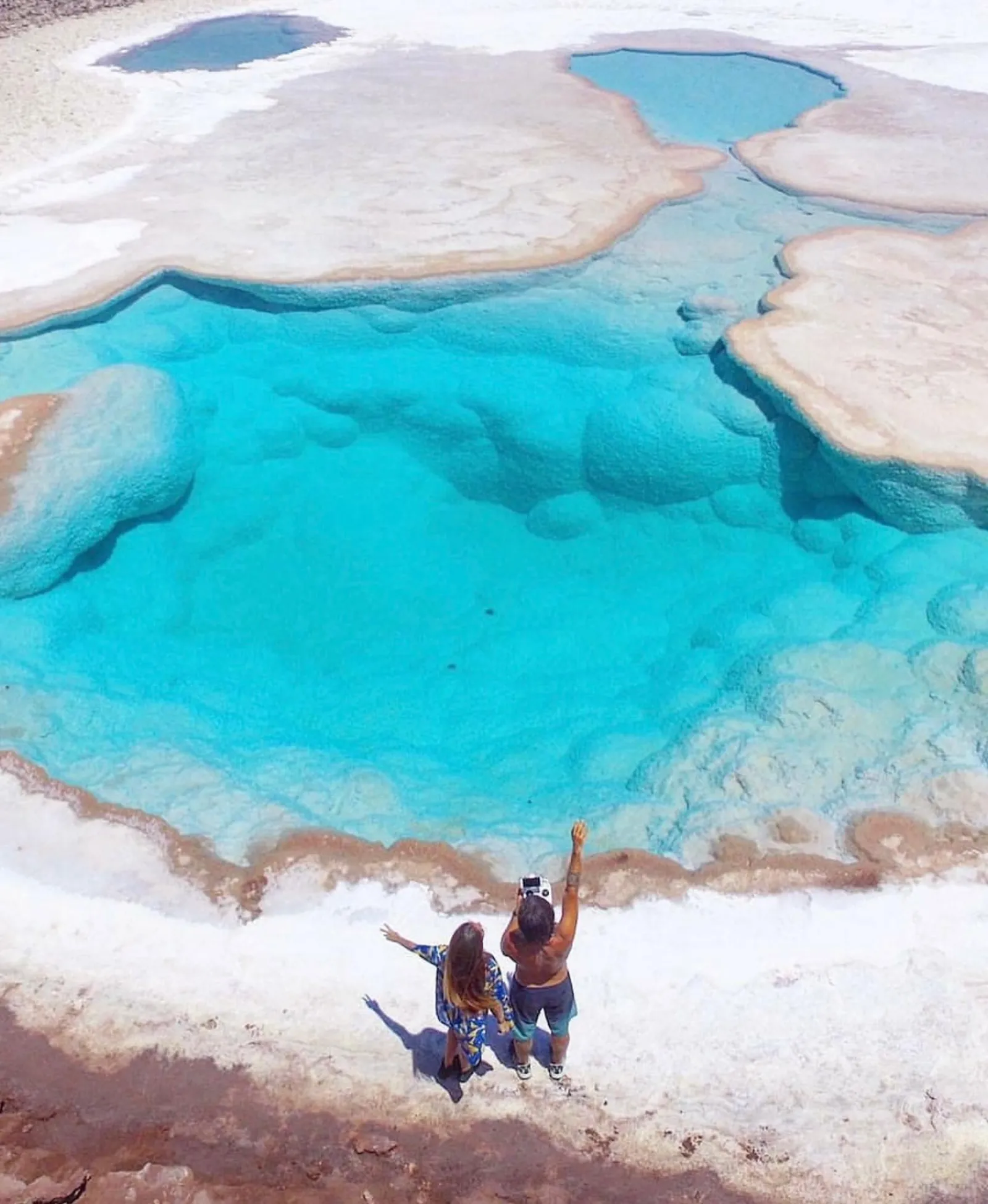 Two people raise their arms at the edge of a bright turquoise lagoon ringed with salt