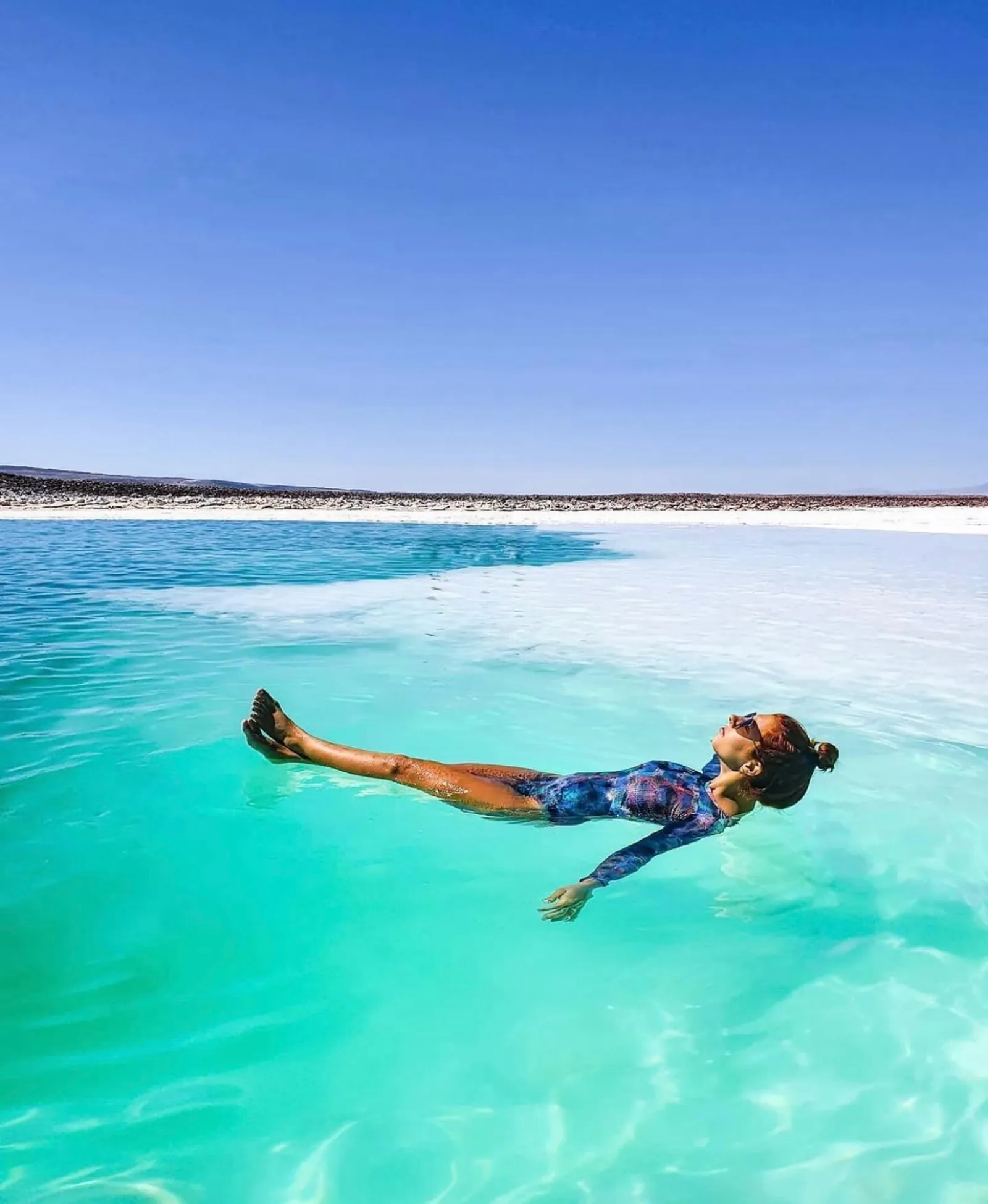 A woman floats in clear turquoise water under a blue sky at a salt lagoon
