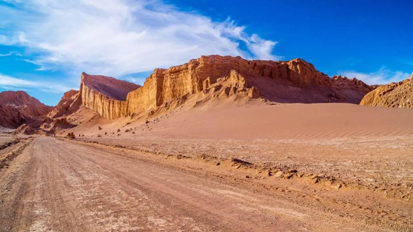 A dirt road passes beneath tall red cliffs and a sand dune in Valle de la Luna