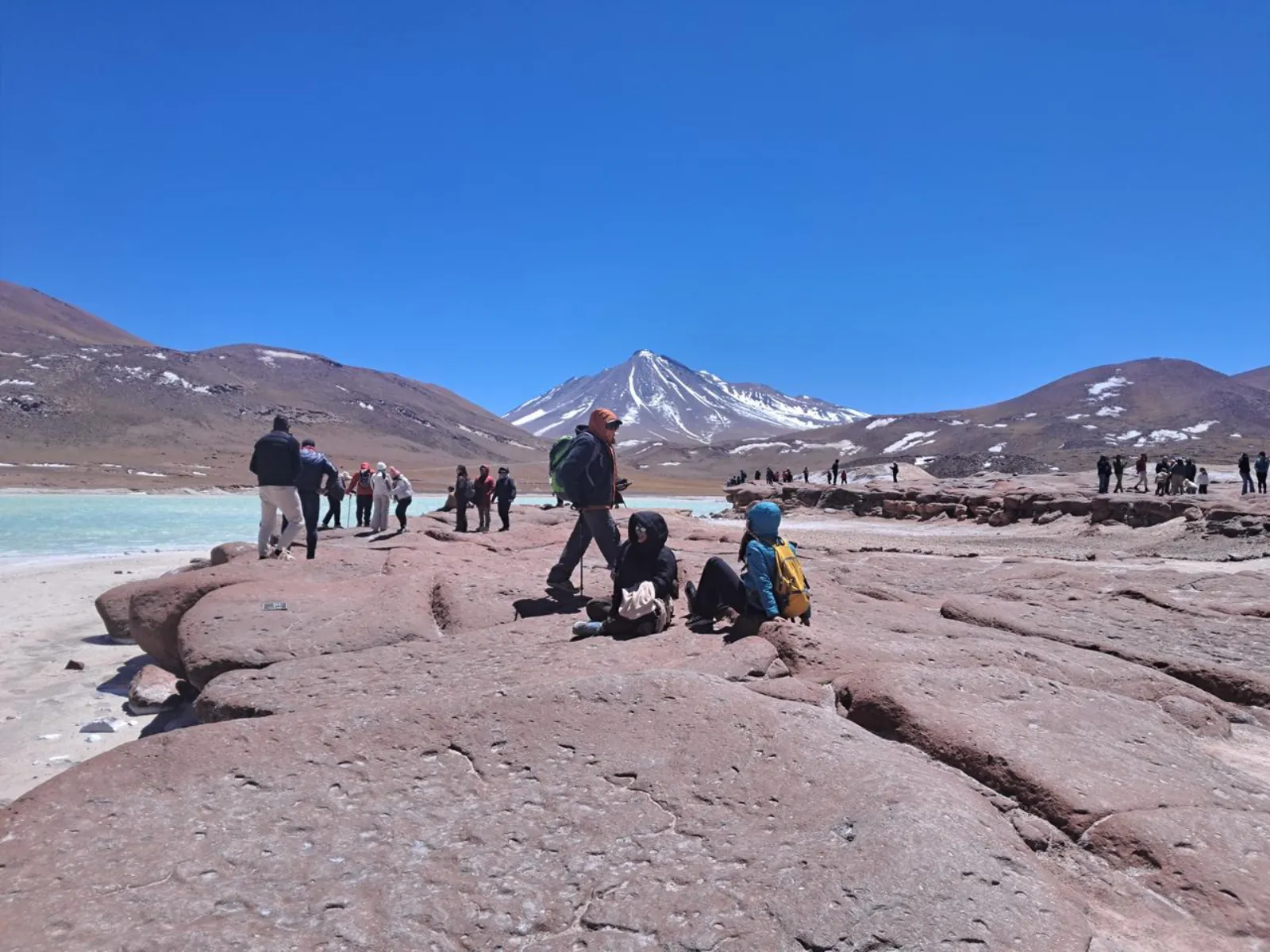 Visitors sit and stand on the red rocks of Piedras Rojas below a snowy volcano