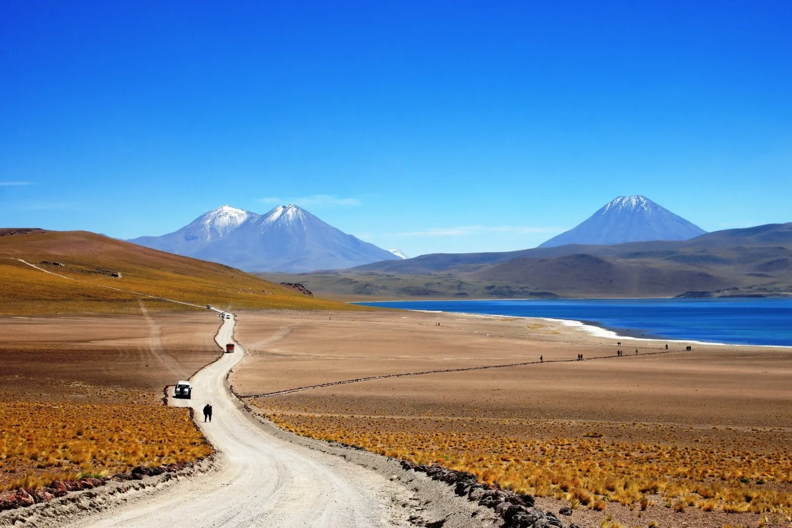 A dirt road leads past a deep blue altiplanic lagoon toward snow-capped volcanoes