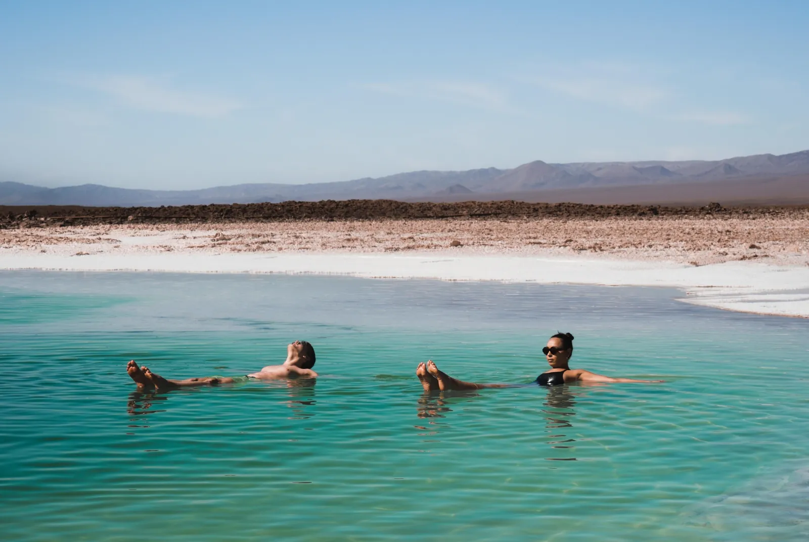 Two travellers float on their backs in a turquoise salt lagoon at Baltinache