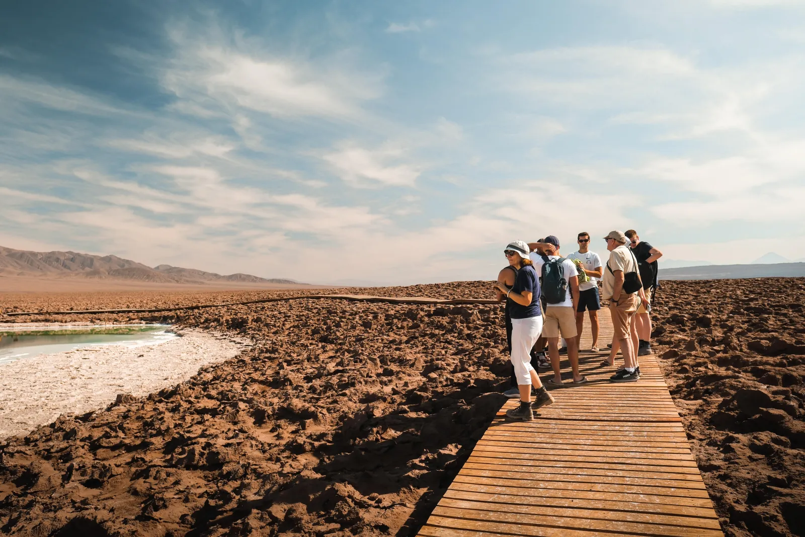 A tour group walks along a wooden boardwalk beside a salt lagoon in the desert