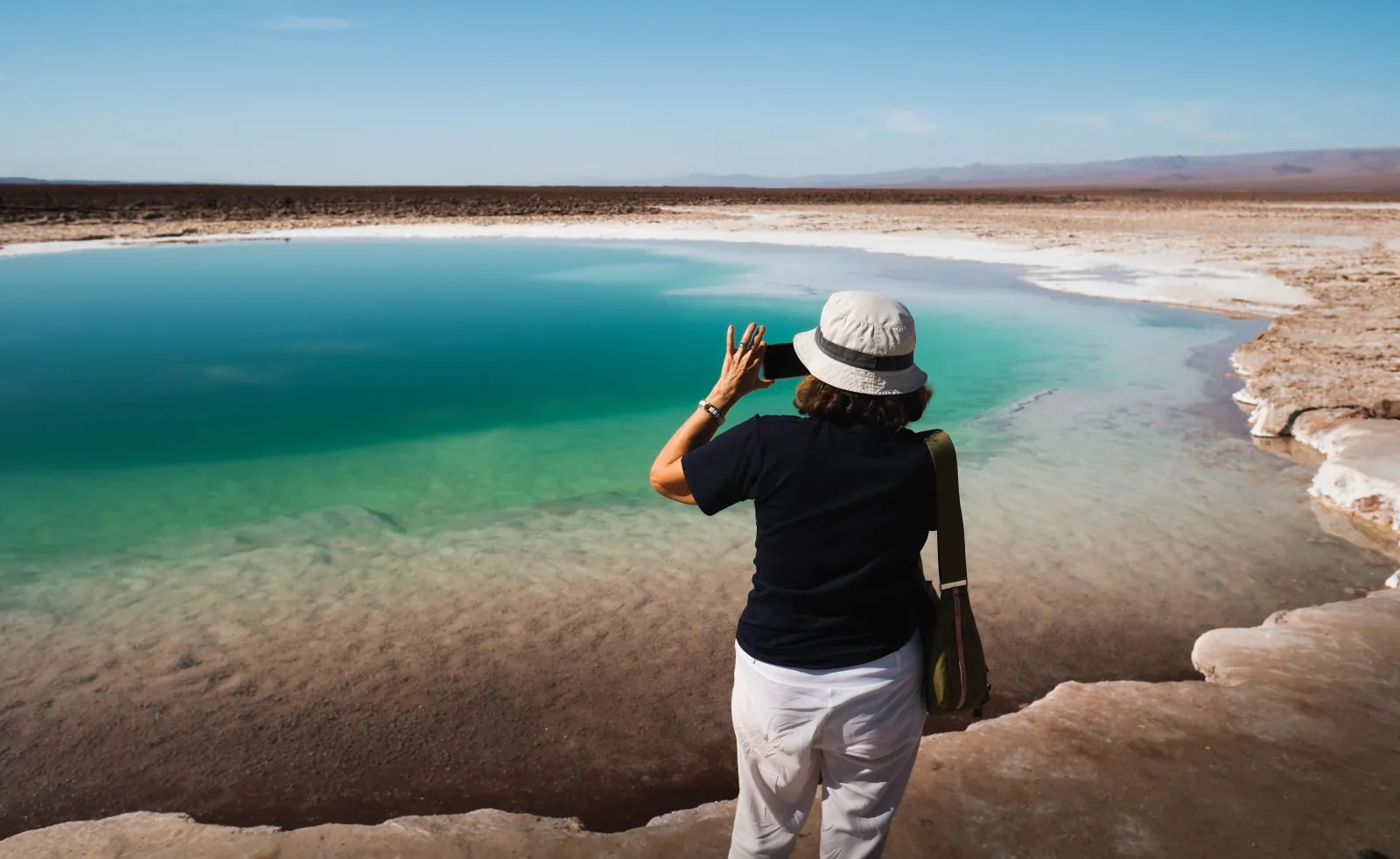 A visitor in a sun hat photographs a turquoise salt-rimmed lagoon