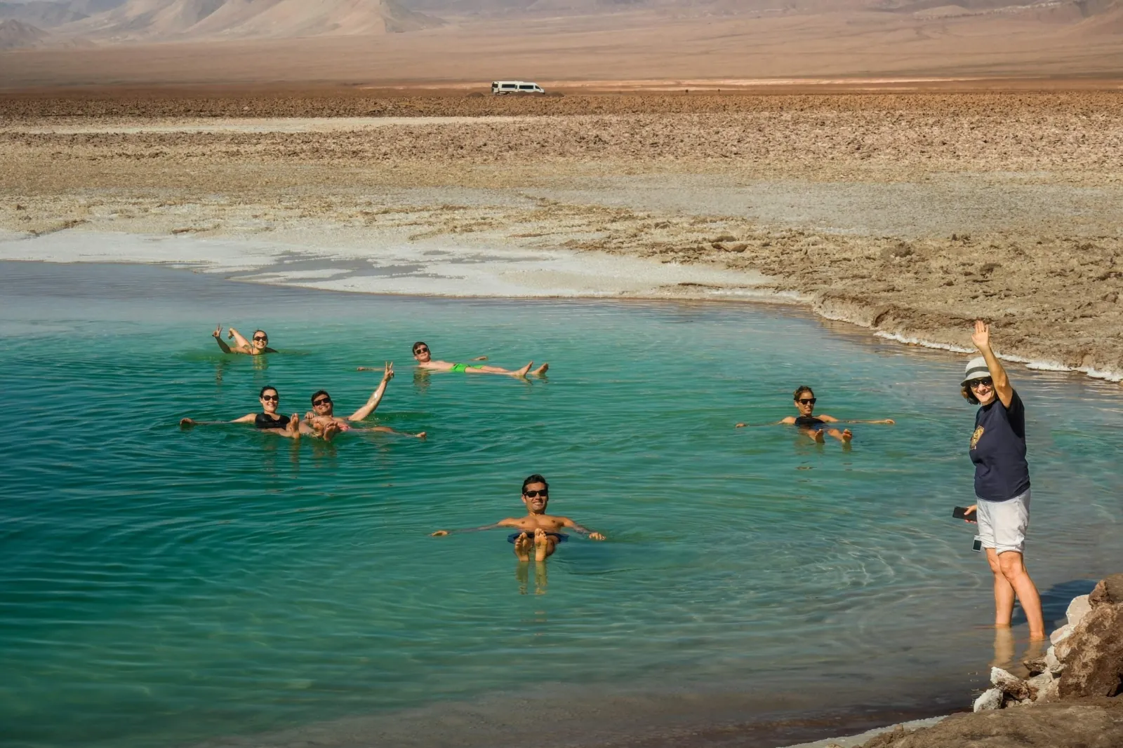 Swimmers float in a green lagoon while a guide waves from the shore, with a van parked beyond