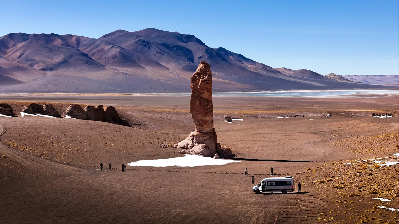 Visitors and a white van below a rock pillar on the plain, with a salt flat and mountains beyond