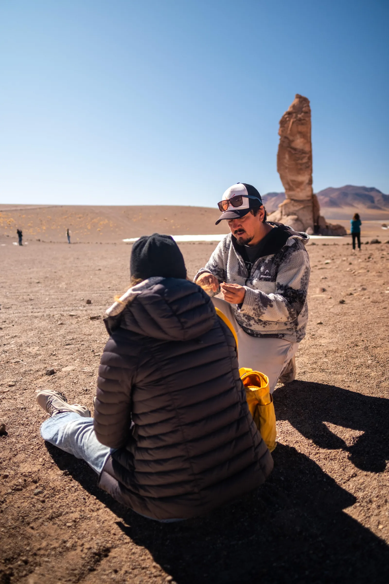 A guide crouches to talk with a seated traveller near the Pacana rock pillars
