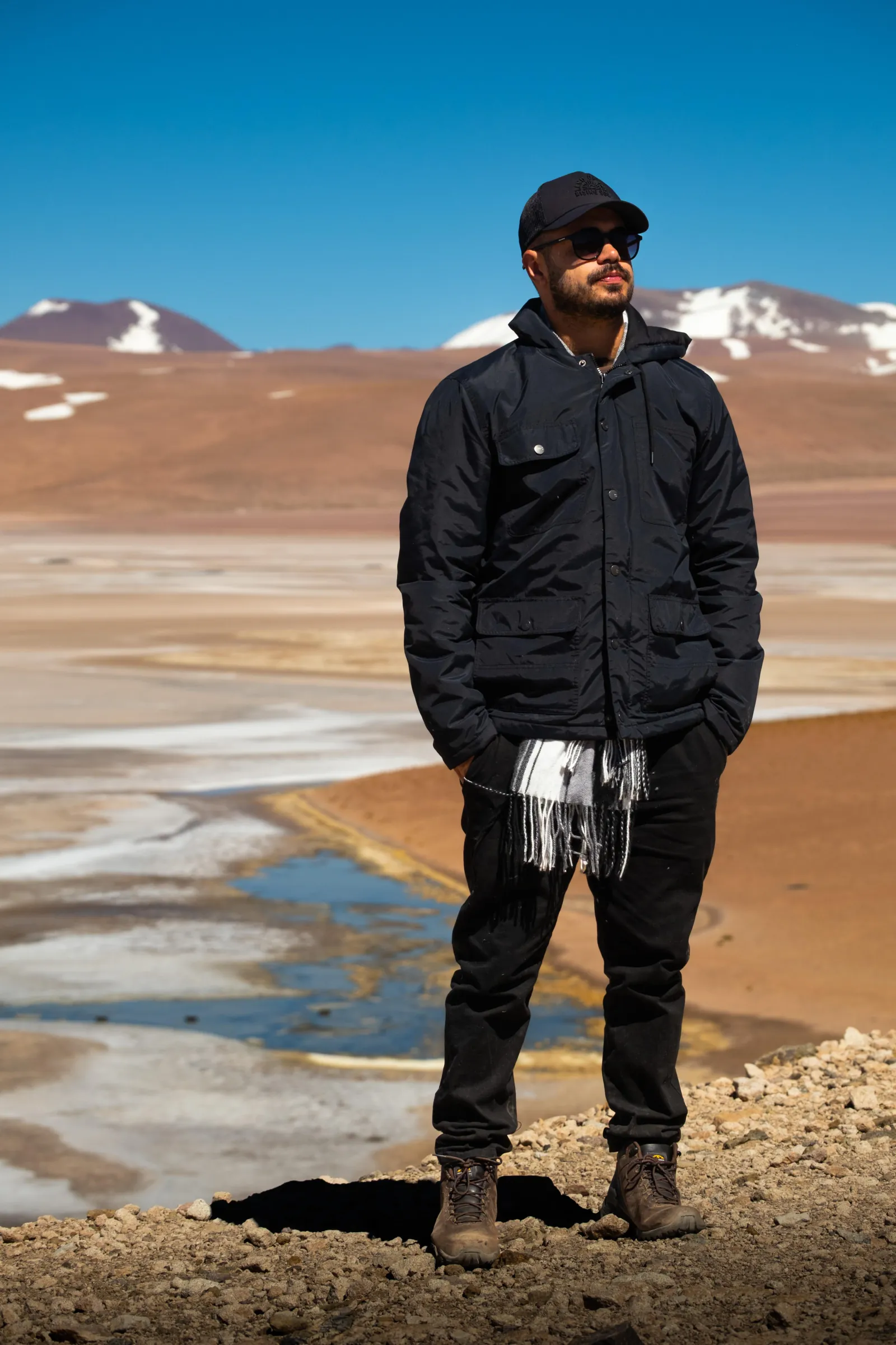 A traveller in a black jacket and scarf stands above a salt flat with water channels