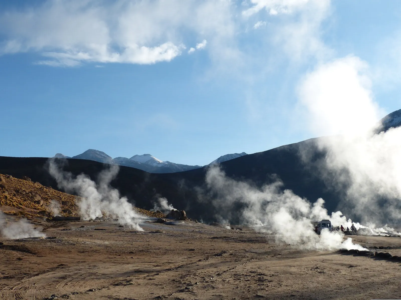 Steam plumes rise across El Tatio, with a vehicle and visitors in the distance