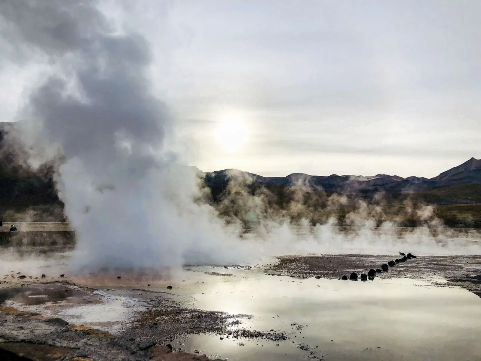 A tall steam column rises over a reflective pool at El Tatio, with the sun behind clouds