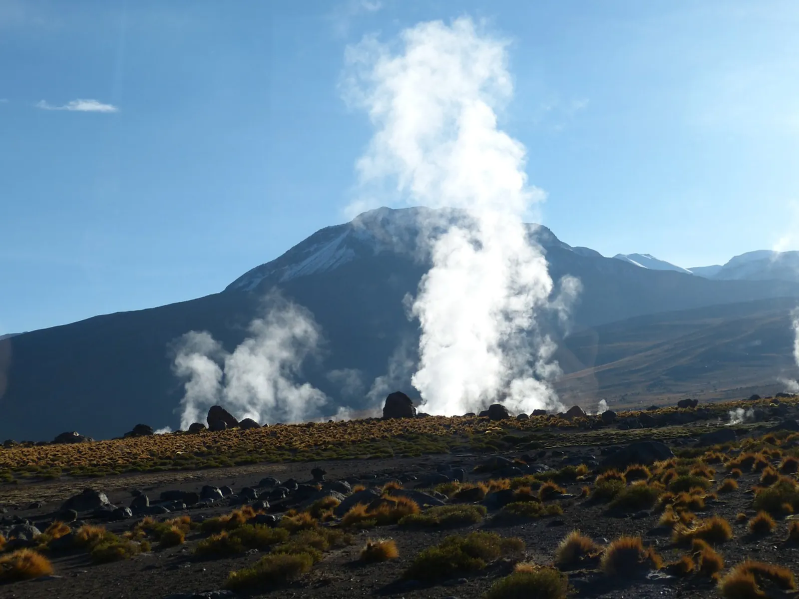 A bright white steam column rises against a dark volcano at El Tatio
