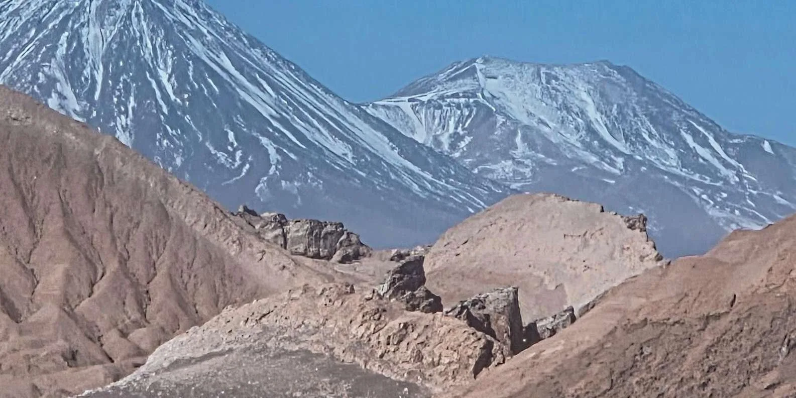 Snow-streaked volcanoes rise behind grey rock ridges seen from Valle de la Luna