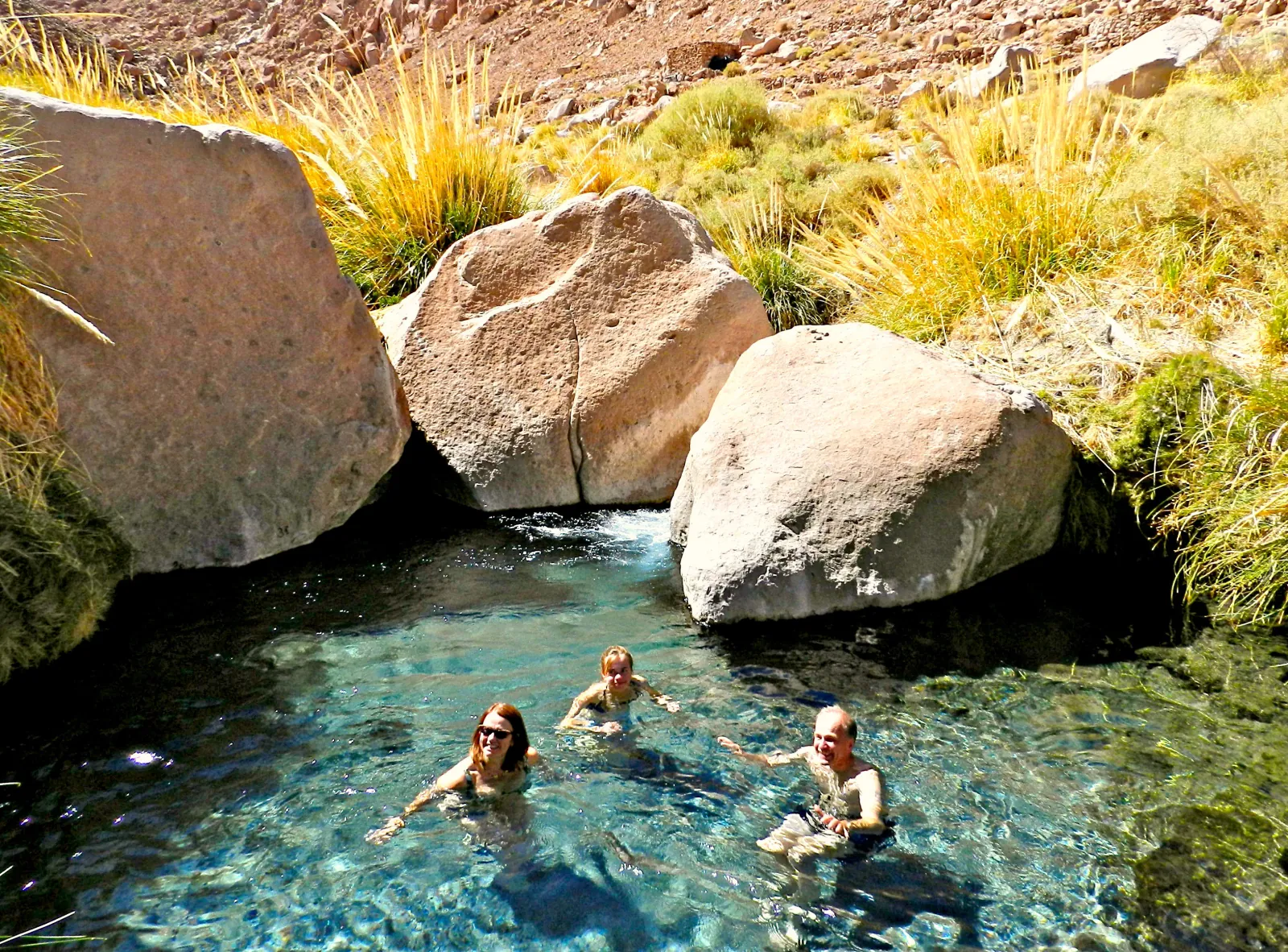 Three bathers in a clear blue-green pool surrounded by boulders and tall grasses
