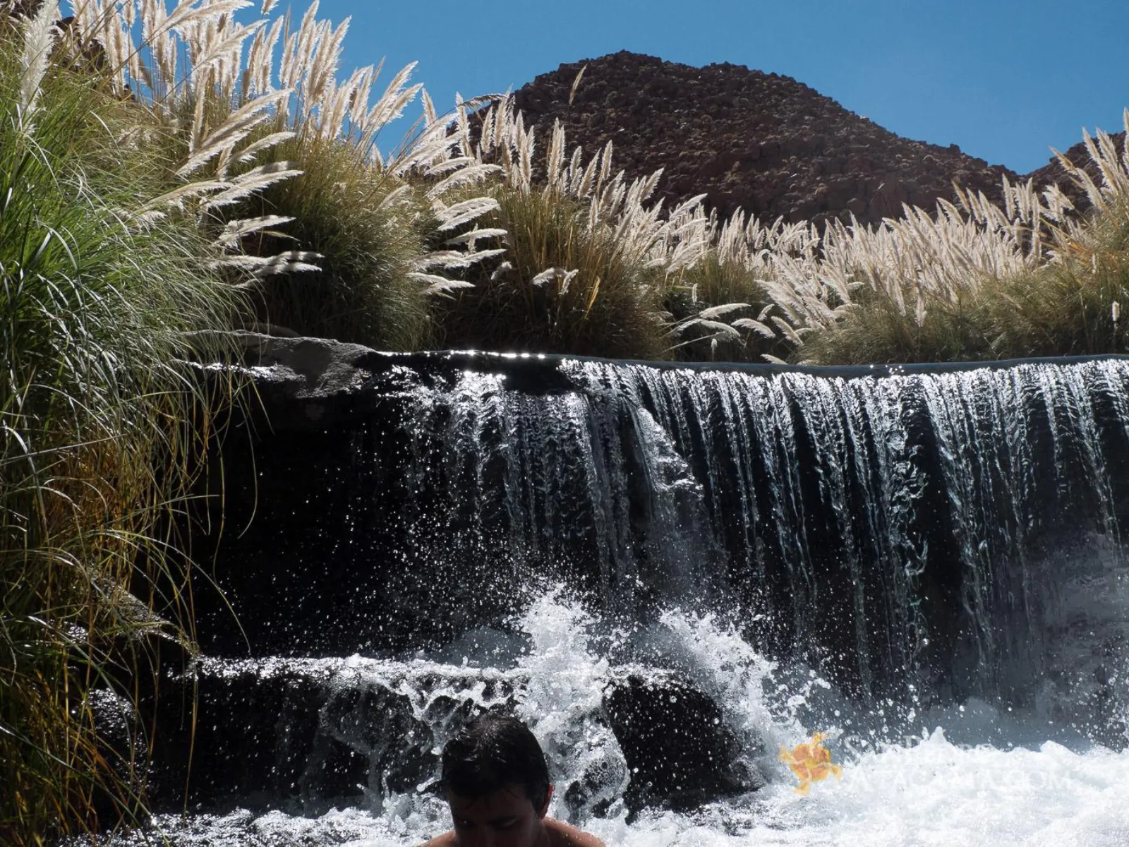 A waterfall pours over a stone ledge into a pool, with a bather in the foreground