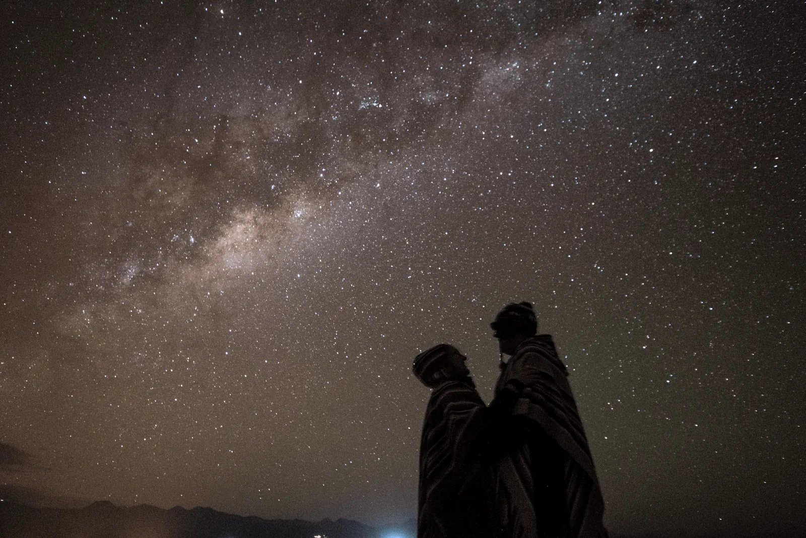 Two guests in hooded ponchos stand in silhouette below a dense field of stars and the Milky Way