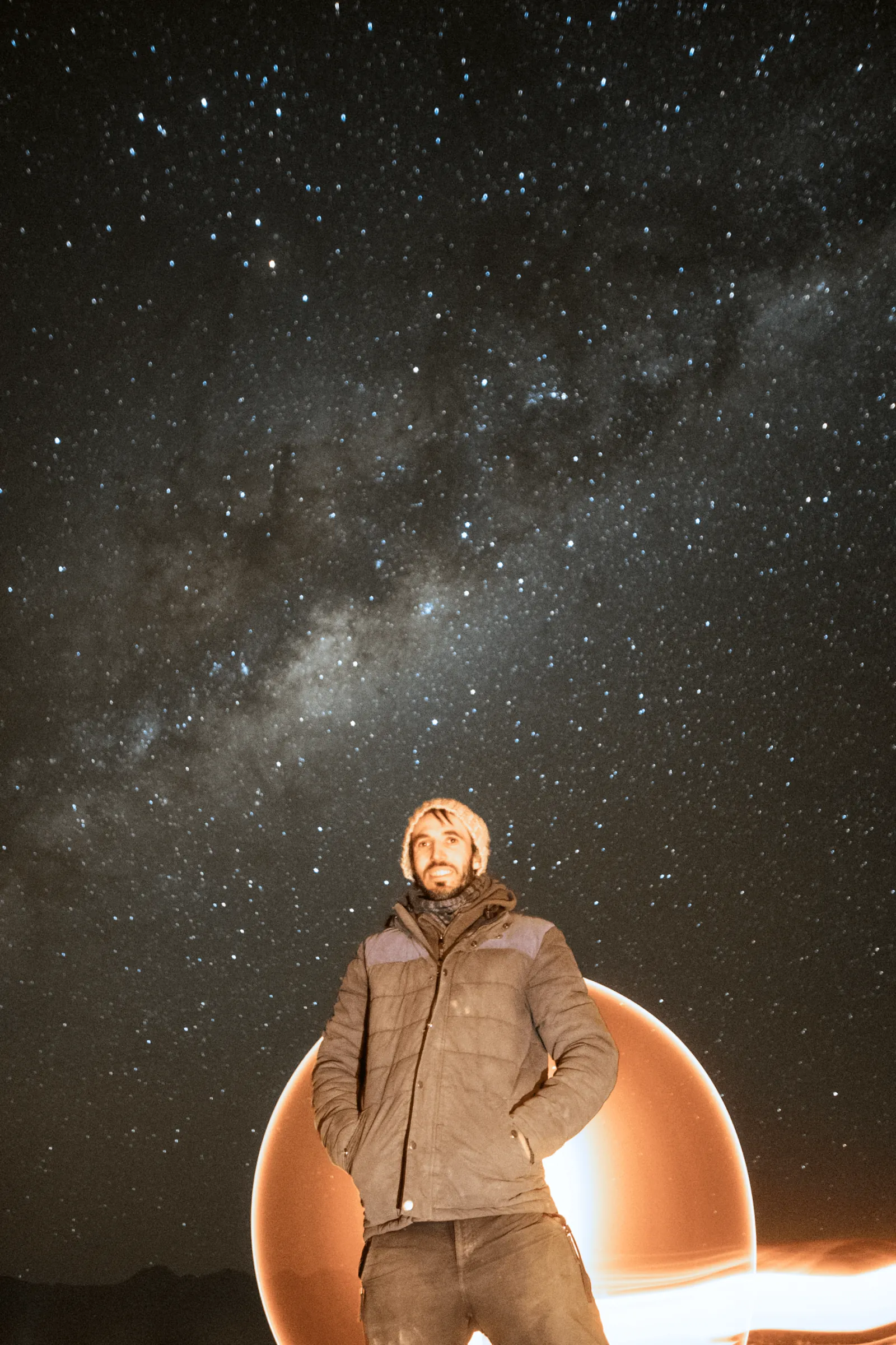A guest in a warm jacket and beanie stands before a glowing light-painted circle under the Milky Way