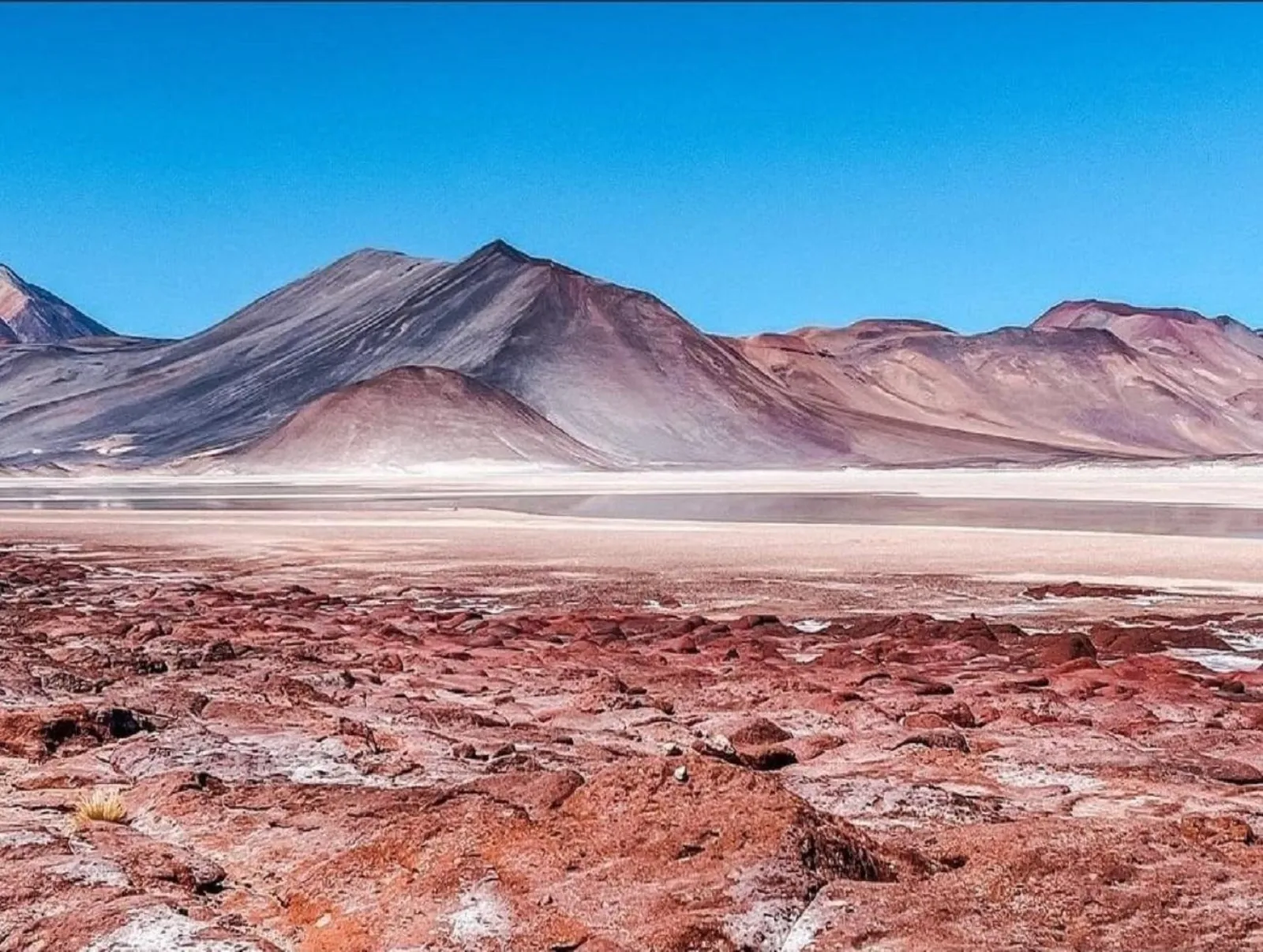 Red rocky ground and a pale salt flat below rounded volcanic mountains in the high Atacama