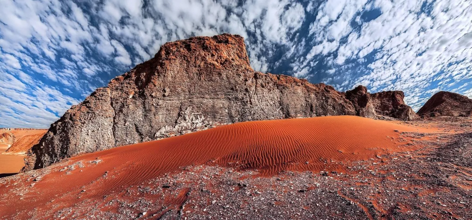 A red sand dune curves below a dark rock outcrop under a mottled sky in the Atacama Desert