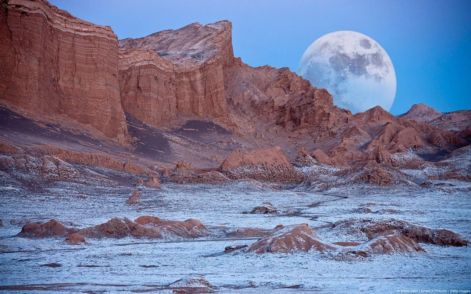 A full moon rises over layered red cliffs and salt-dusted ground in the Atacama Desert