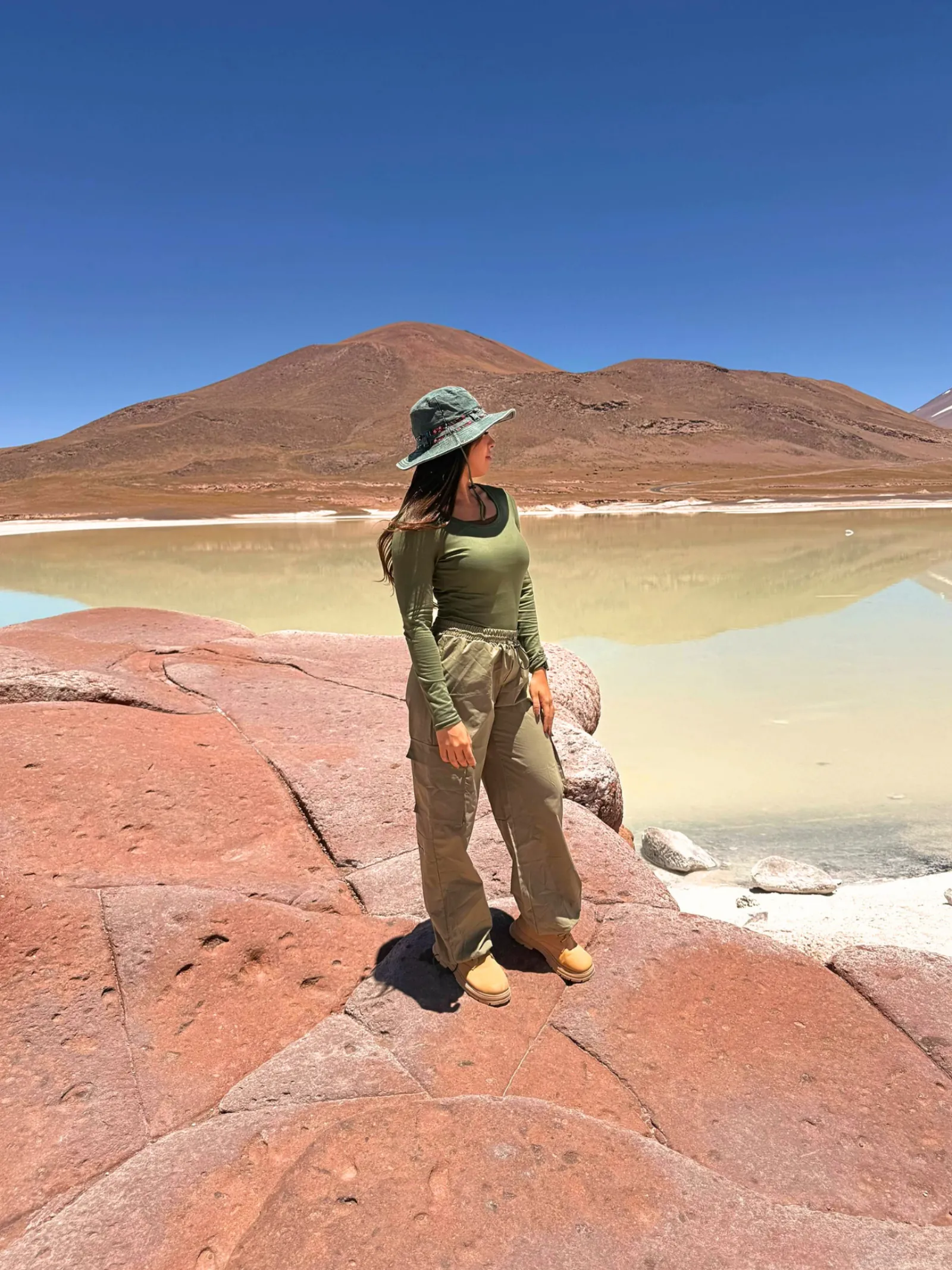 A traveller in a sun hat stands on red rock at Piedras Rojas beside a still salt lagoon
