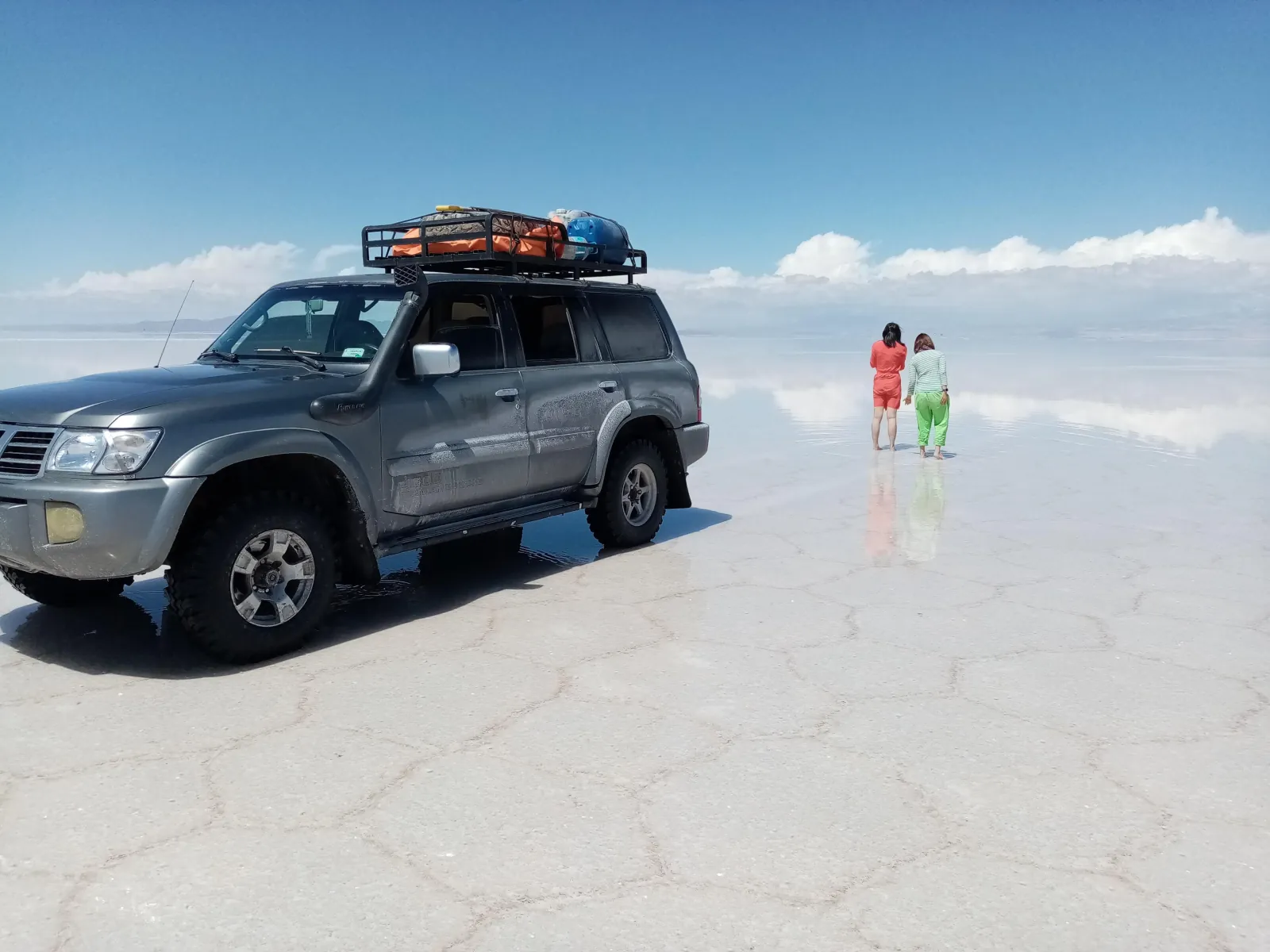 A 4x4 with a loaded roof rack on the wet Uyuni salt flat as two travellers walk across the reflection