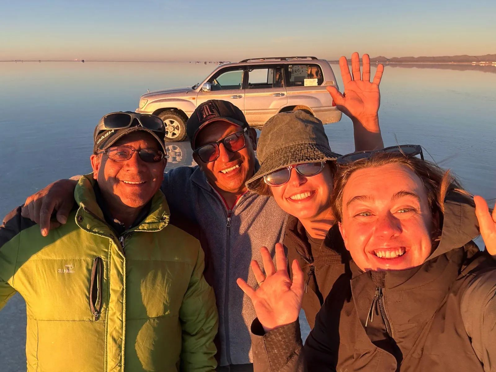 Smiling travellers wave for a selfie in front of their 4x4 on the Uyuni salt flat