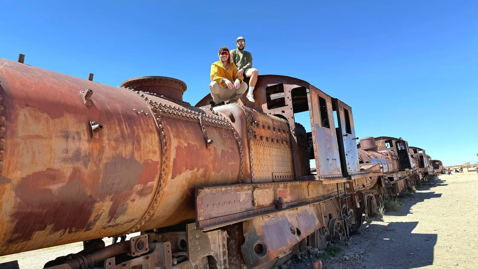 Two travellers sit on top of a rusting steam locomotive at the Uyuni train cemetery