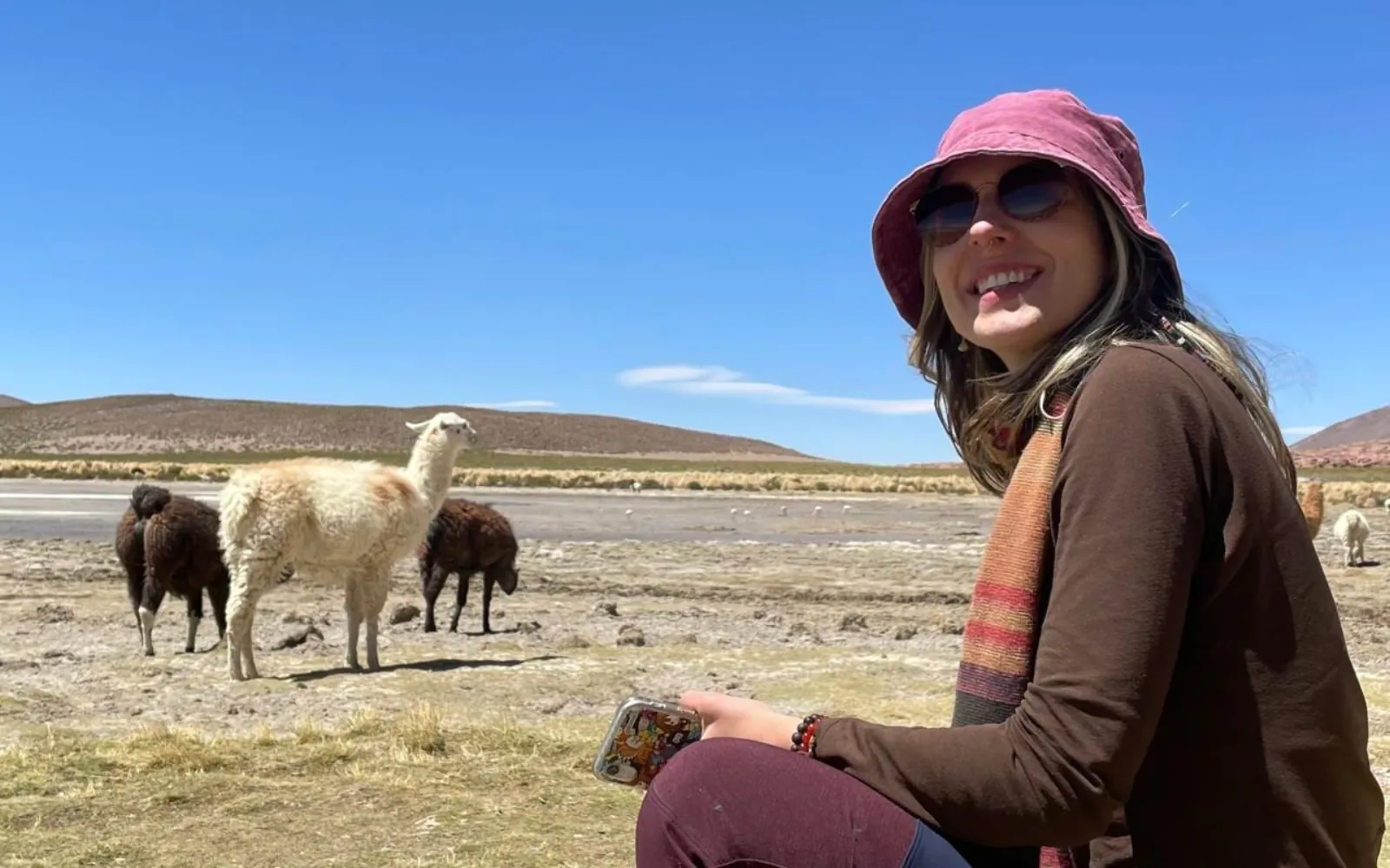 A smiling traveller in a bucket hat sits on the altiplano with llamas behind her