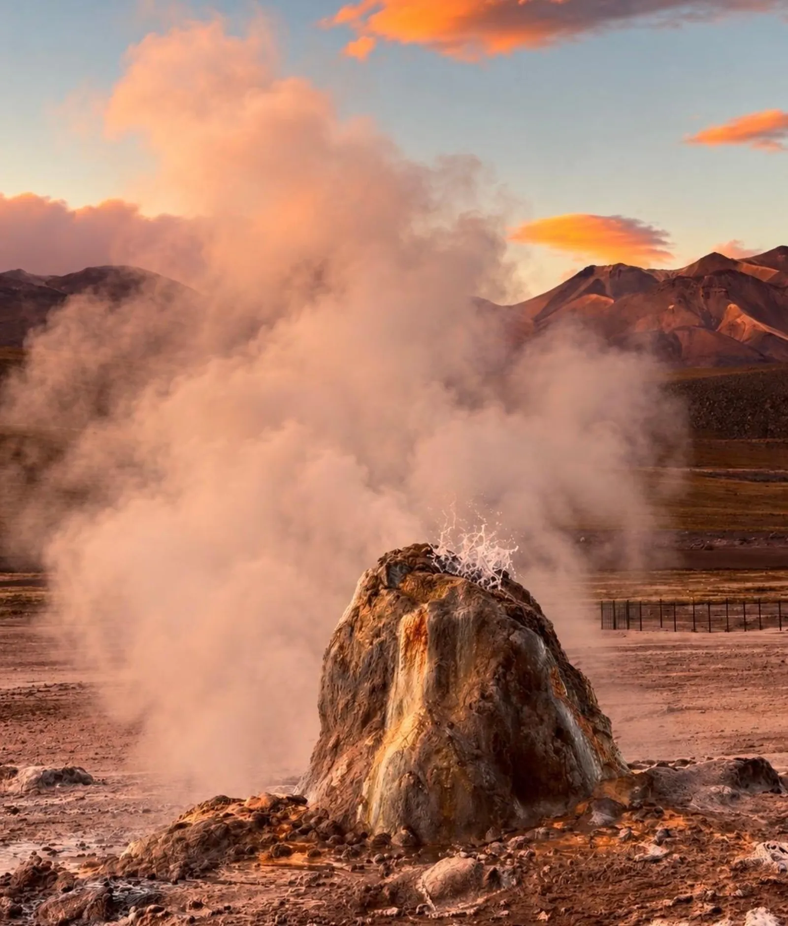 Steam drifts from a mineral cone at El Tatio as sunrise colours the clouds and mountains