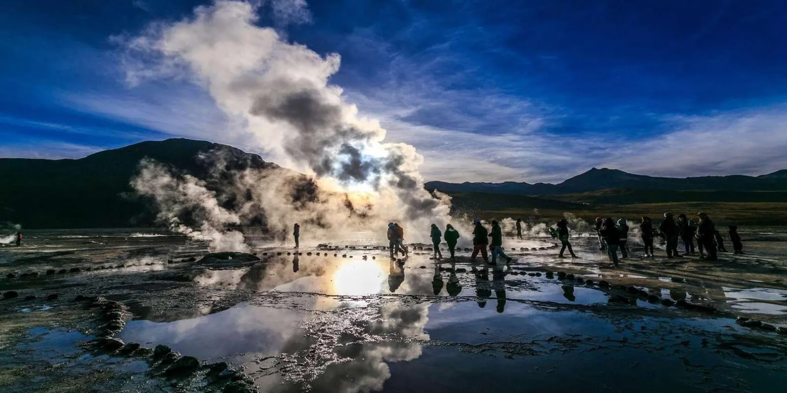 Visitors walk among steaming vents and reflective pools at El Tatio under a deep blue sky