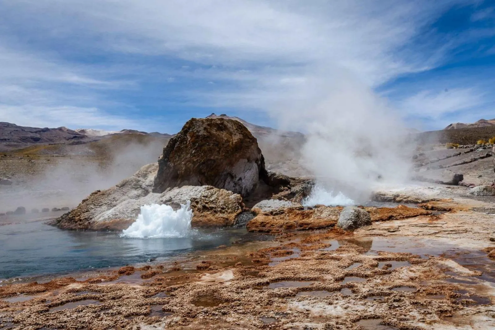 Boiling water bursts from beside a rock mound at El Tatio, with steam rising behind